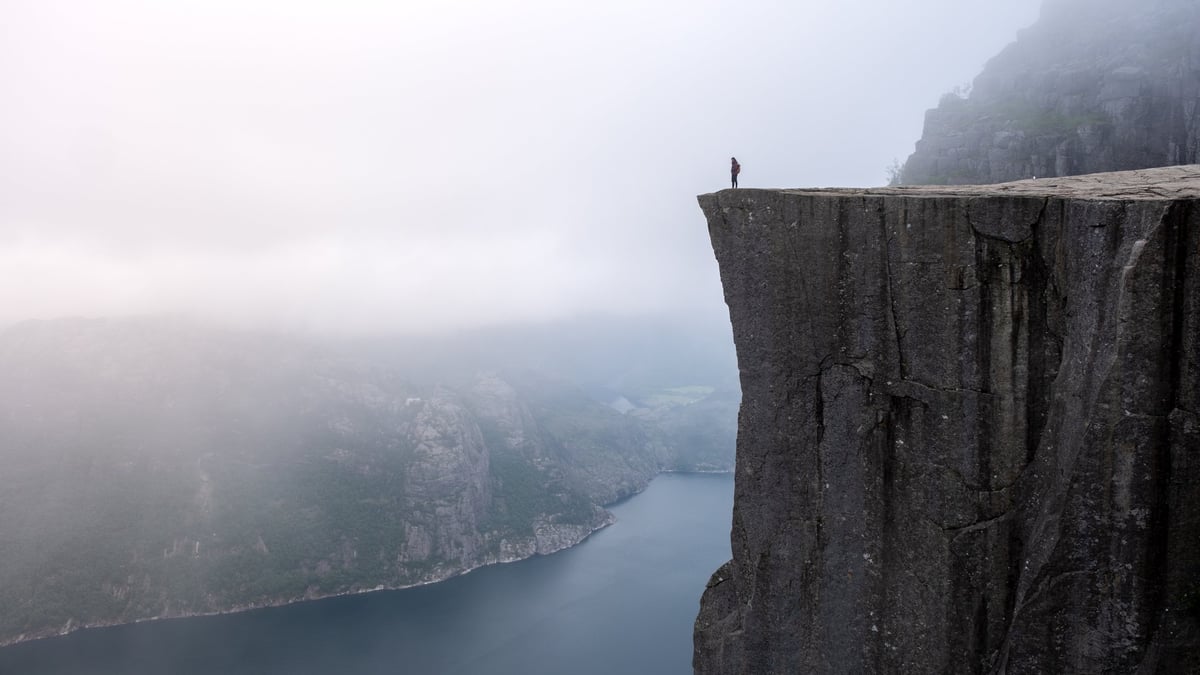 A solitary figure stands at the edge of Preikestolen, gazing over the misty Lysefjord as dawn breaks, casting ethereal light on the dramatic cliffs and tranquil waters below.